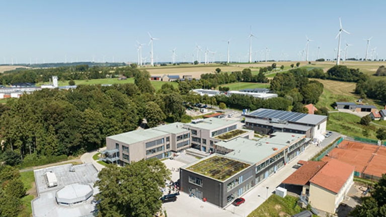 Moderne Campusgebäude mit begrünten Dächern und Windrädern im Hintergrund.