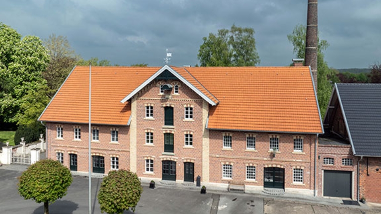 Backsteinbau mit rotem Ziegeldach und hohem Schornstein, eingerahmt von Bäumen.
