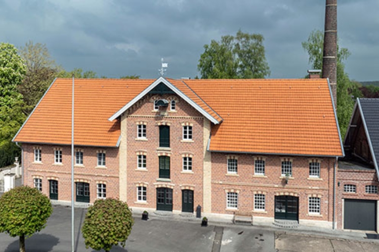 Backsteinbau mit rotem Ziegeldach und hohem Schornstein, eingerahmt von Bäumen.