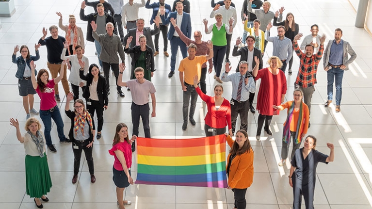 Bunt gekleidete Mitarbeiterinnen und Mitarbeiter der NRW.BANK halten einen Regenbogen-Fahne hoch