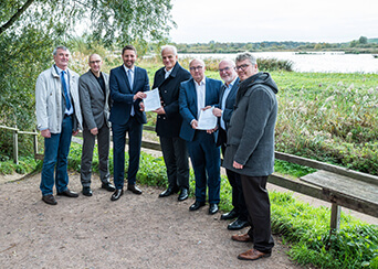 7 Männer bei der Übergabe eines Förderbescheids vor einer Seenlandschaft