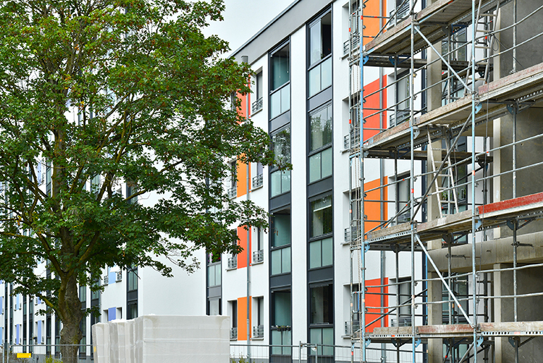 Ein fertiger Gebäudeteil mit weißer Fassade und orangenen Schmuckelementen neben einem eingerüsteten Gebäudeteil.
