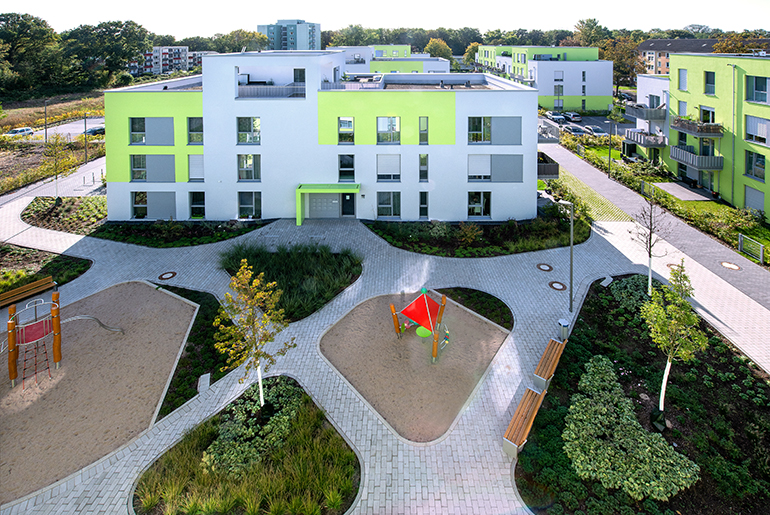 Grün-graue Häuser mit Flachdächern umrahmen einen begrünten Innenhof mit Spielplatz