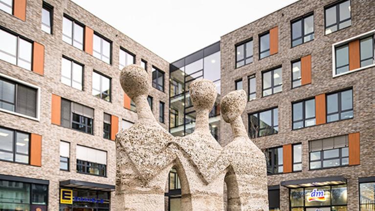 Steinernes Standbild vor einem modernen Gebäudekomplex mit Wohneinheiten, Super- und Drogeriemarkt