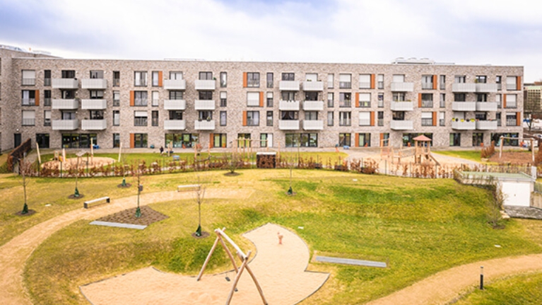 Hausfront einer modernen Wohnanlage, davor eine große Grünfläche mit Spielplatz