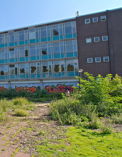 Außenansicht einer ehemaligen Schule mit zerstörten Fenstern, davor eine Brachfläche