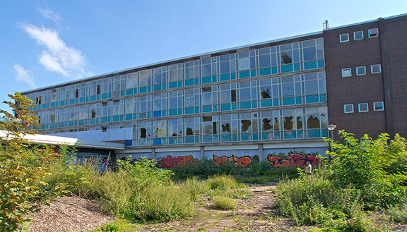 Außenansicht einer ehemaligen Schule mit zerstörten Fenstern, davor eine Brachfläche