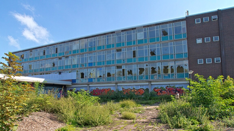 Außenansicht einer ehemaligen Schule mit zerstörten Fenstern, davor eine Brachfläche