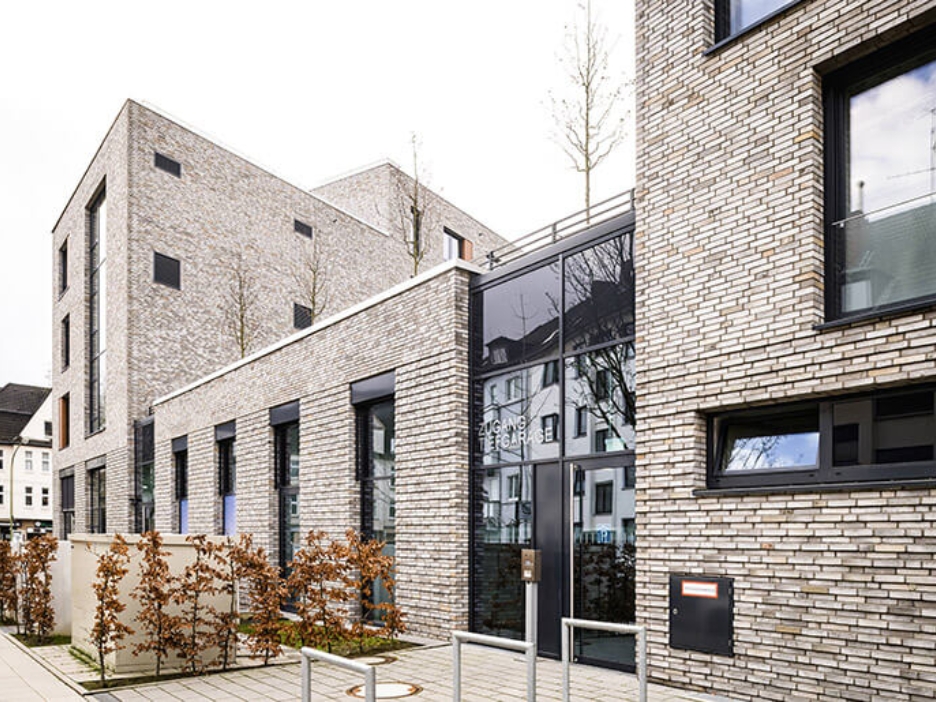 Ein Wohnhaus mit Flachdach und heller Klinkerfassade. Davor und darauf Bäume und Büsche mit herbstlichen Laub.