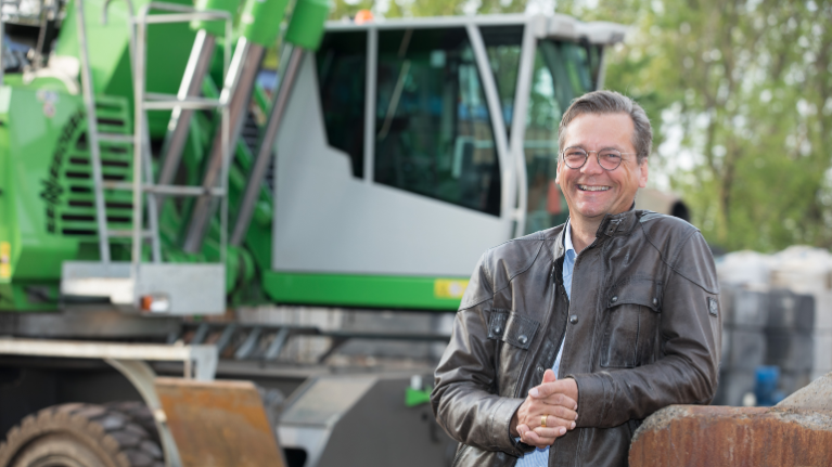Ein Mann in Lederjacke steht vor einer großen Baumaschine und lächelt in die Kamera.