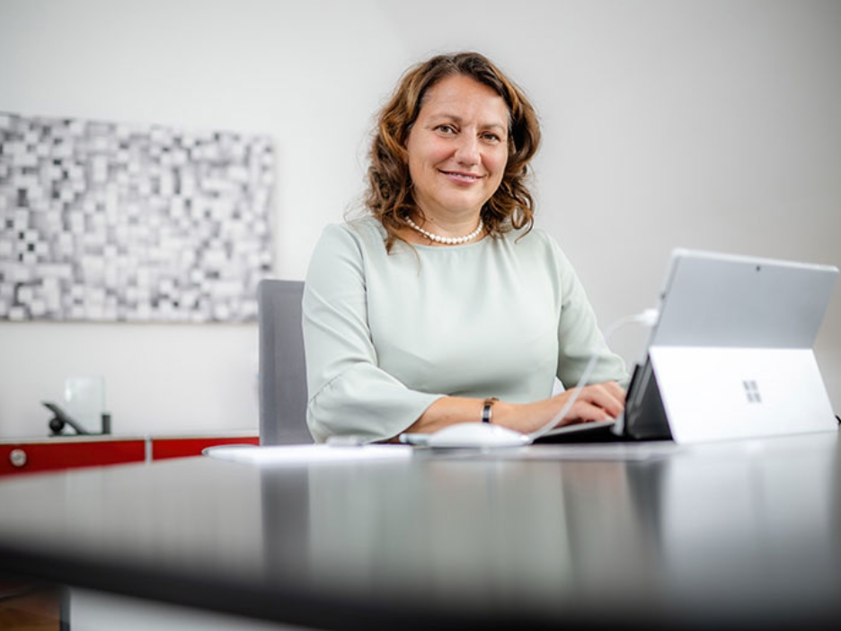 Sabine Hansen sitzt in einem weißen, hellen Büro an ihrem Schreibtisch, tippt etwas in ihr Tablet und schaut in die Kamera.