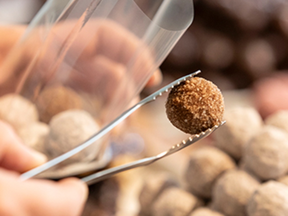 Eine handgemachte Praline wird mit einer Zange in eine Tüte eingepackt. im Hintergrund sind weitere Pralinen zu sehen.