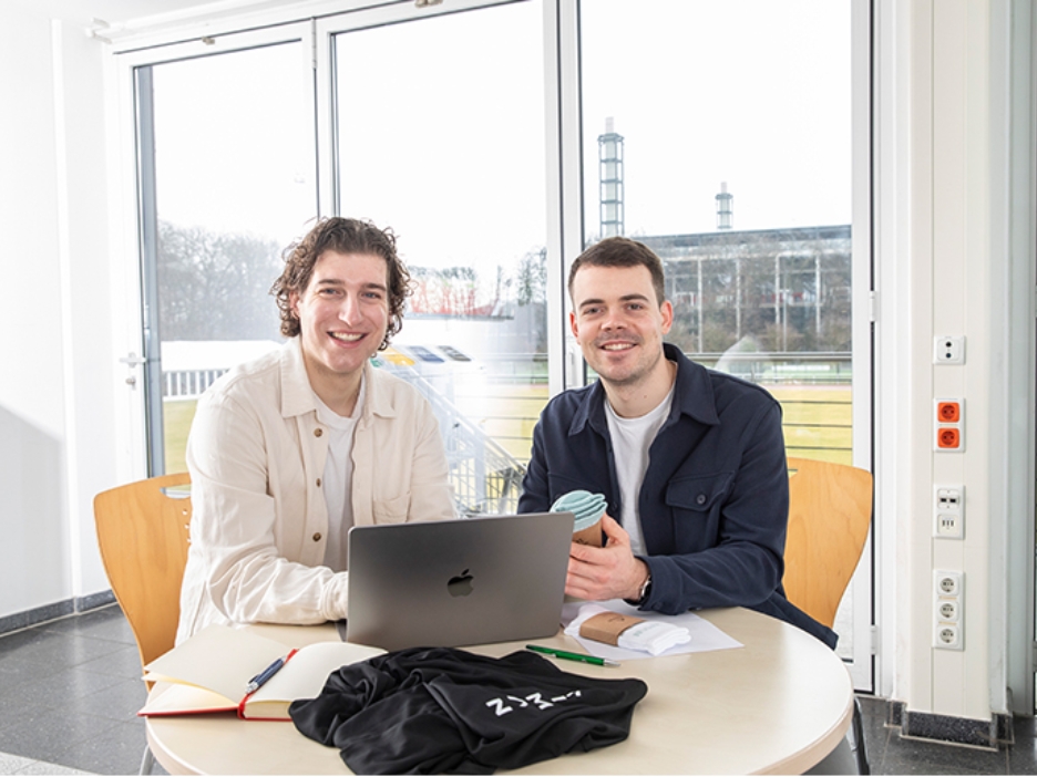 Die zwei GEschäftsführer von Kiman Sport sitzen an einem Laptop und halten Produkte ihrer Sportartikel Firma in den Händen