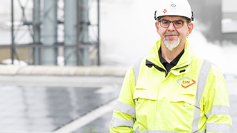 Andreas Fedderwitz, Elektroingenieur bei dem Recyclingbetrieb, steht in gelber Warnjacke auf einer Anhöhe vor den Fabrikdächern mit Solarmodulen