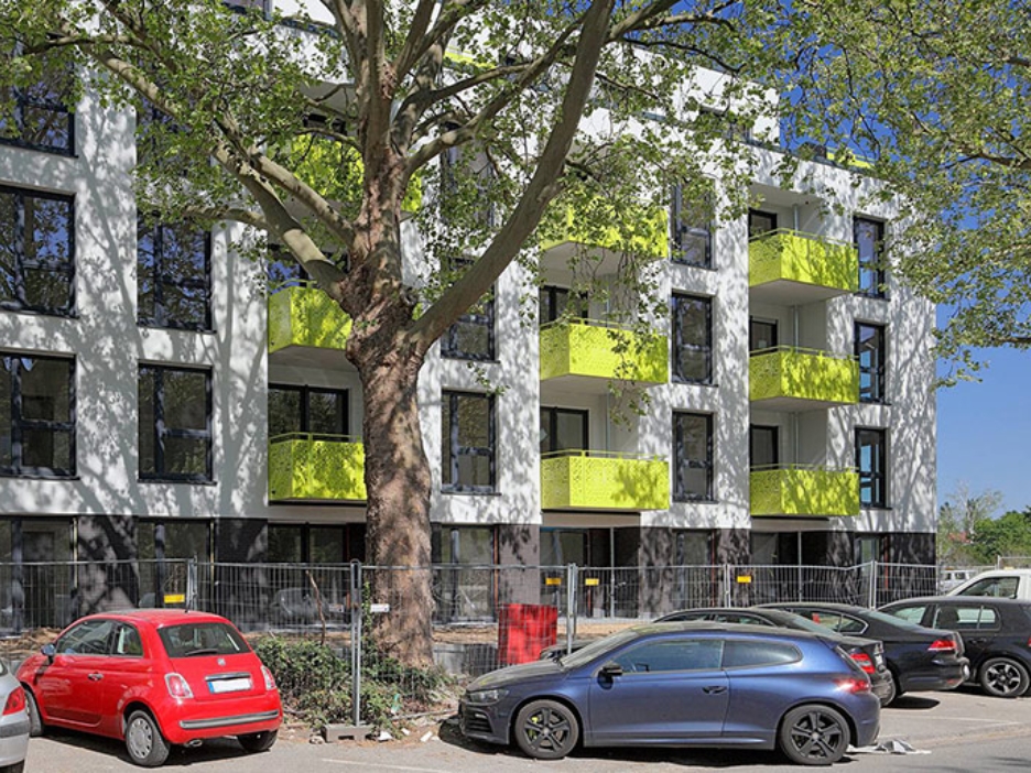 Weißes Mehrfamilienhaus mit gelben Balkonen, davor Bäume, ein Bauzaun und parkende Autos