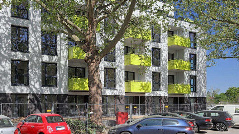 Weißes Mehrfamilienhaus mit gelben Balkonen, davor Bäume, ein Bauzaun und parkende Autos