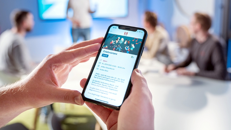 Das Foto zeigt Hände, die ein Smartphone halten, auf dem die Oberfläche der App Tremaze zu finden ist. Im Hintergrund sind vier Männer zu sehen, die ein Meeting abhalten.