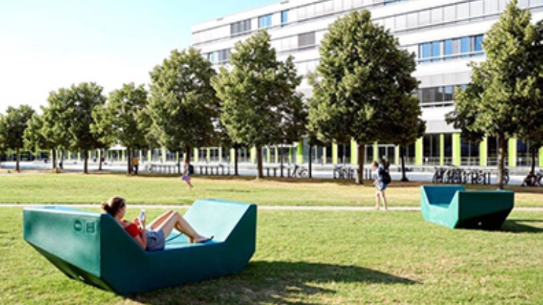 Wiese mit Bäumen und Studierenden, im Hintergrund ein Gebäude der Universität Bielefeld