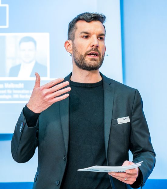 Ein Mann im dunklen Anzug spricht auf einer Bühne und gestikuliert mit einer Hand, während er Moderationskarten in der anderen hält.