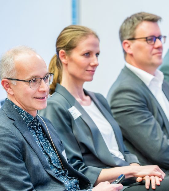 Drei Personen in Businesskleidung sitzen nebeneinander auf Stühlen und hören aufmerksam zu.
