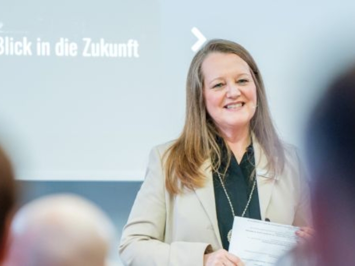 Eine Frau mit Mikrofon spricht vor Publikum, im Hintergrund ist der Schriftzug „Blick in die Zukunft“ zu sehen.