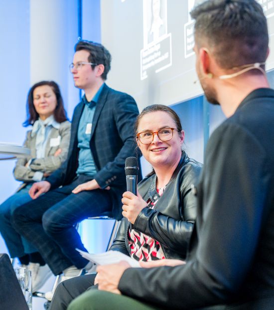 Vier Personen sitzen auf einer Bühne, eine Frau mit Mikrofon spricht, während die anderen zuhören.